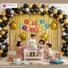 Birthday party setup with DECORATIONkart’s 66 pcs decoration kit featuring a multicolor cardstock “Happy Birthday” banner, golden fringe curtains, metallic balloons in golden, black, and silver, golden star foils, and DIY accessories.