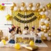 Birthday party setup with DECORATIONkart’s 66 pcs decoration kit featuring a black cardstock “Happy Birthday” banner, golden fringe curtains, metallic balloons in golden, yellow, and white, golden star foils, and DIY accessories.