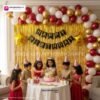 Birthday party setup with DECORATIONkart’s 66 pcs decoration kit featuring a black cardstock “Happy Birthday” banner, golden fringe curtains, metallic balloons in golden, red, and white, golden star foils, and DIY accessories.