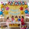 Birthday party setup with DECORATIONkart’s 66 pcs decoration kit featuring a blue cardstock “Happy Birthday” banner, golden fringe curtains, multicolor metallic balloons, star foils in red, blue, golden, and pink, and DIY accessories.