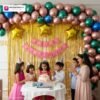 Birthday party setup with DECORATIONkart’s 66 pcs decoration kit featuring a pink cardstock “Happy Birthday” banner, golden fringe curtains, multicolor metallic balloons, star foils in red, blue, golden, and pink, and DIY accessories.
