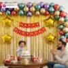 Birthday party setup with DECORATIONkart’s 66 pcs decoration kit featuring a red cardstock “Happy Birthday” banner, golden fringe curtains, multicolor metallic balloons, star foils in red, golden, pink, and blue, and DIY accessories.