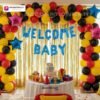 Welcome baby party setup with DECORATIONkart’s 64 pcs decoration kit featuring a blue dotted foil “Welcome Baby” banner, golden fringe curtains, metallic balloons in yellow, red, and black, multicolor star foils, and DIY accessories.